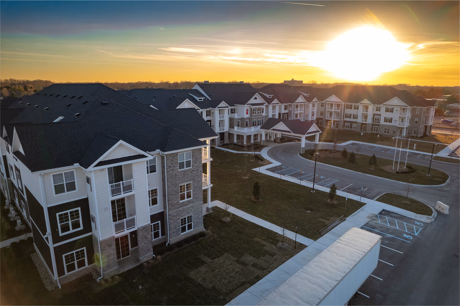 Aerial view of a large senior living facility building with multiple floors, surrounded by parking spaces and landscaped grounds, captured during sunset with the sun low on the horizon.