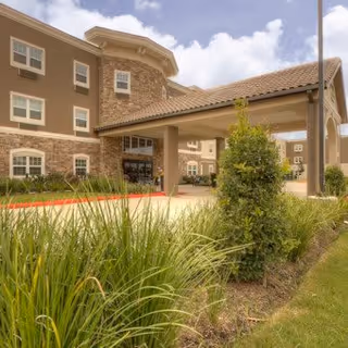 Exterior view of a senior living facility building with a covered entrance, stone and beige siding, multiple windows, and landscaped greenery including bushes and grass under a partly cloudy sky.
