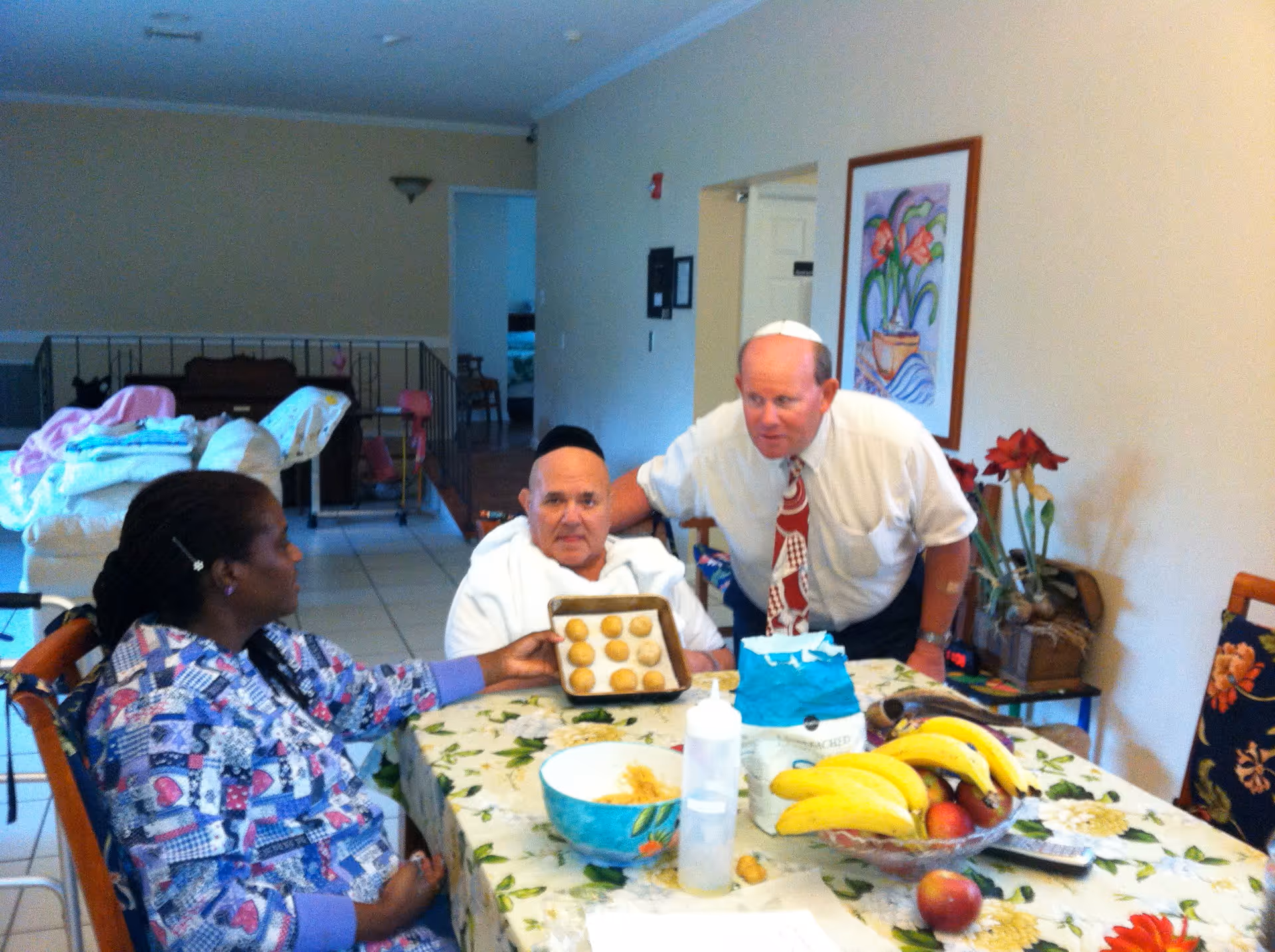 Three people sitting around a dining table in a room. One person is holding a baking tray with round dough balls. The table has a floral tablecloth, a bowl, a bottle, a bag of flour, and a bowl of bananas and apples. The room has light-colored walls, a framed flower painting, and some furniture in the background.