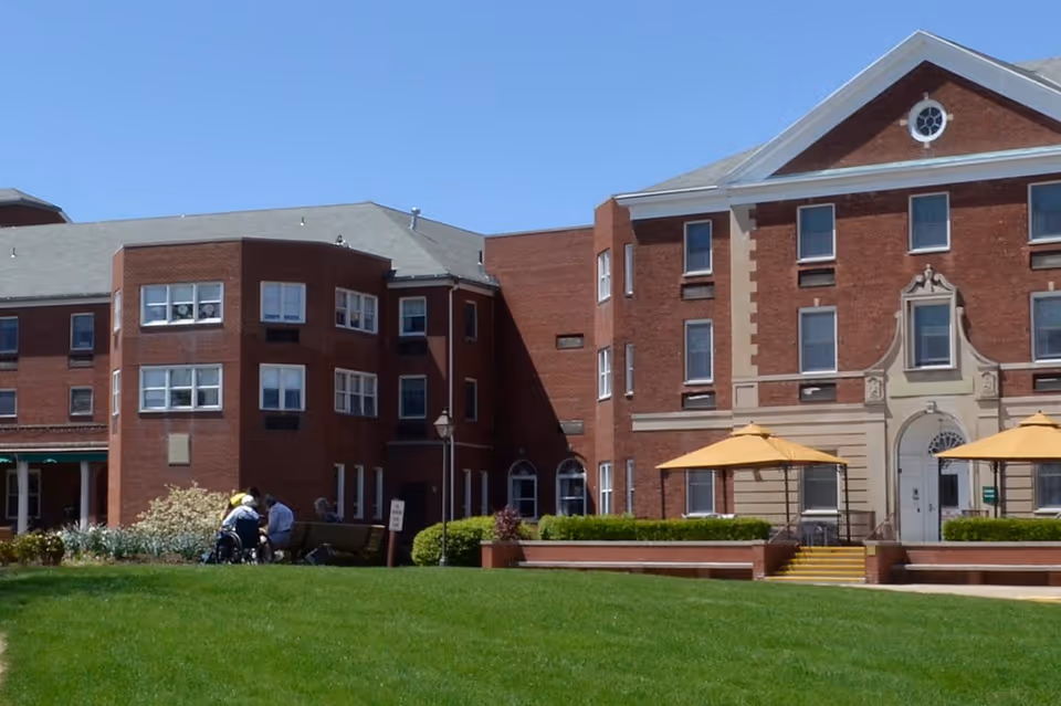 Exterior view of a large brick building with multiple windows and a central entrance with steps. There are two yellow umbrellas shading outdoor seating areas on a raised patio. In the foreground, a green lawn is visible with a few people sitting on benches near the building.