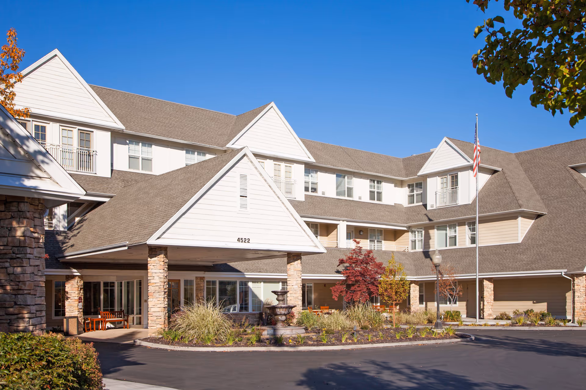 Exterior view of The Wellington senior living facility showing a large building with multiple windows, a covered entrance supported by stone pillars, landscaped plants, a fountain, and an American flag on a flagpole under a clear blue sky.