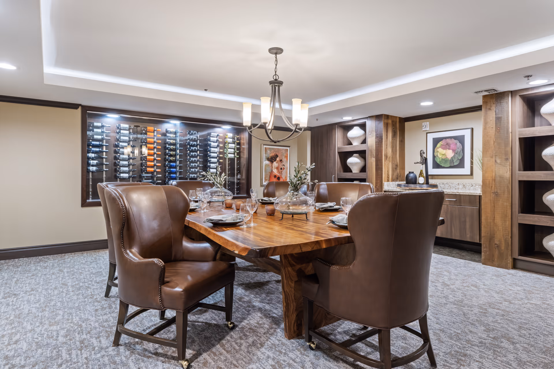 A well-lit dining room with a large wooden table surrounded by six brown leather chairs. The table is set with plates, glasses, and decorative centerpieces. Behind the table is a wall-mounted wine rack filled with bottles, and built-in wooden shelves with decorative vases. The room has a modern chandelier hanging from the ceiling and framed artwork on the walls.