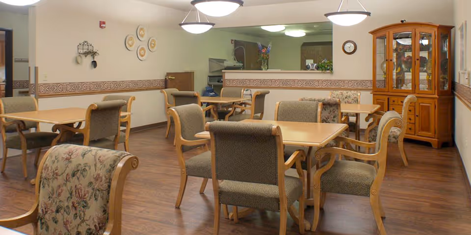 A dining room in a senior living facility with several wooden tables and cushioned chairs arranged neatly. The room has wood flooring, beige walls with a decorative border, and ceiling lights. A wooden china cabinet with glass doors is visible on the right side, and a pass-through window to the kitchen area is in the background.