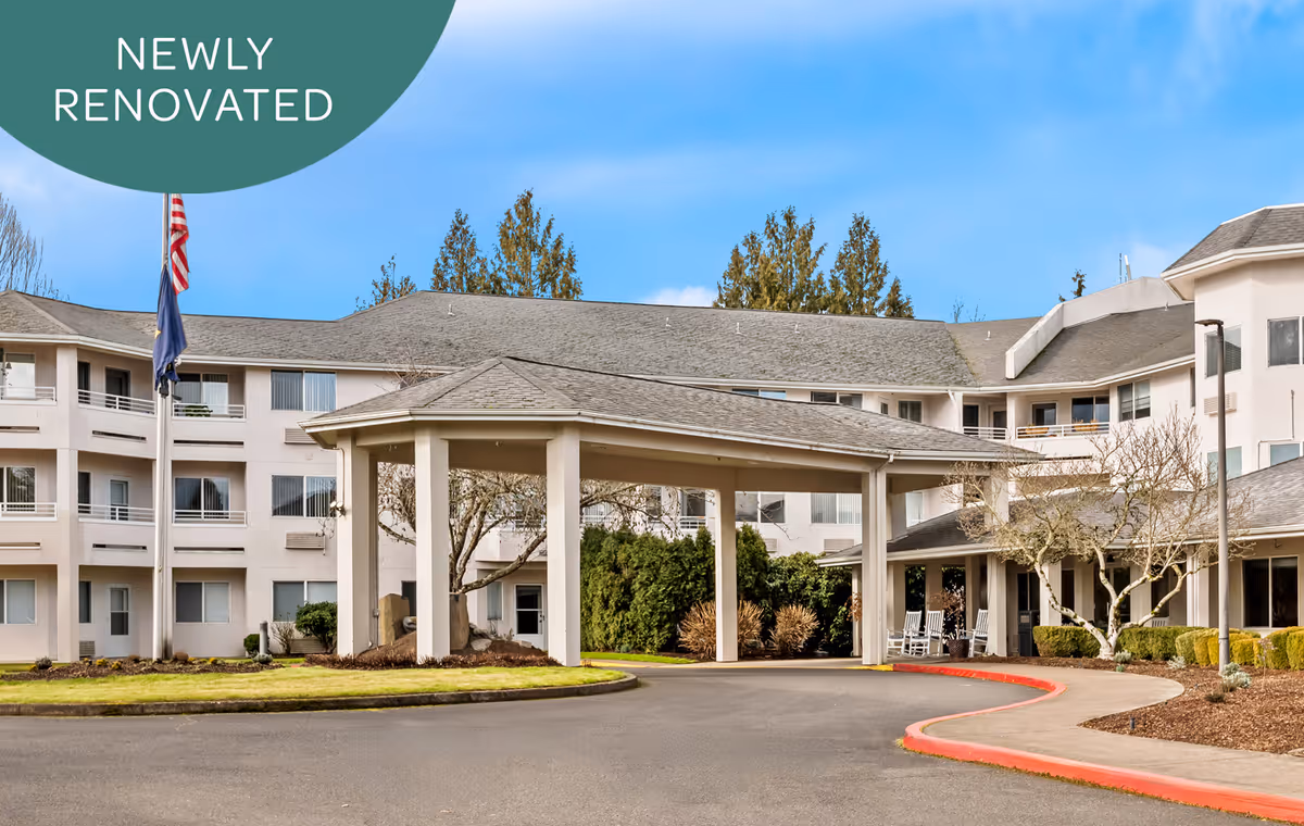 Exterior view of a large, newly renovated senior living facility building with a covered entrance, multiple windows, and a driveway. There are two flagpoles with flags, some trees, and landscaping around the building under a clear blue sky.