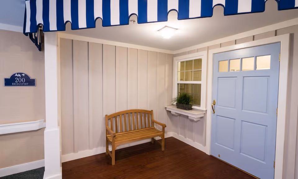 An indoor entryway area with a light blue door featuring four small windows at the top. Next to the door is a small window with a white shelf underneath holding a potted plant. A wooden bench is placed against a beige paneled wall. Above, a blue and white striped awning extends across the ceiling. A sign on the left wall reads '200 Bridgeport'. The floor is wooden.