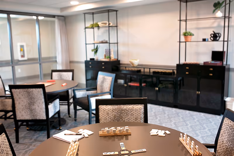 A well-lit common room with round tables and cushioned chairs arranged around them. On the closest table, there is a game of dominoes set up with tiles and racks. In the background, there are black cabinets and shelves with decorative plants and items. Large windows with curtains allow natural light into the room.