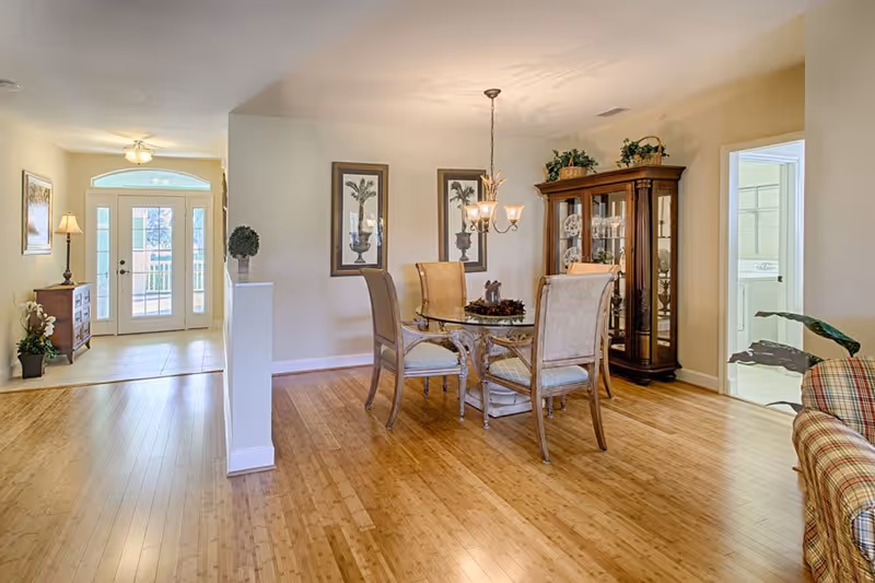 A bright dining area with a round glass table and four cushioned chairs on a wooden floor. Two framed palm tree pictures hang on the wall behind the table. A wooden china cabinet with glass doors is positioned in the corner, decorated with plants and dishes. To the left, there is an entryway with double glass doors and a small wooden chest with a lamp and plant. To the right, a doorway leads to a laundry room with a washer and dryer visible. Part of a plaid upholstered chair and a large green plant are visible in the foreground.