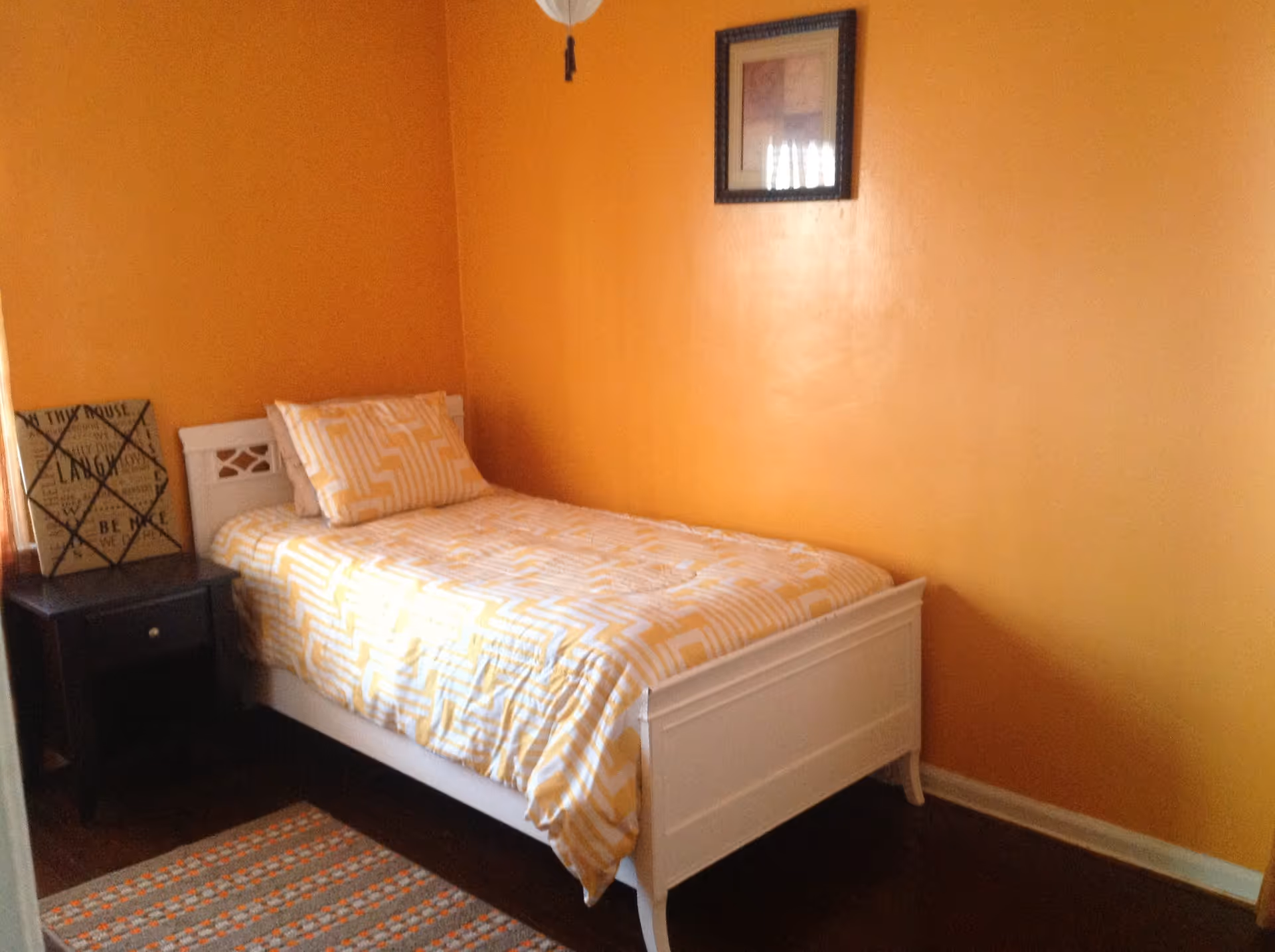 A small bedroom with orange walls featuring a single white bed with yellow and white patterned bedding. Next to the bed is a dark wooden nightstand with a decorative sign on it. A framed picture hangs on the wall above the bed.