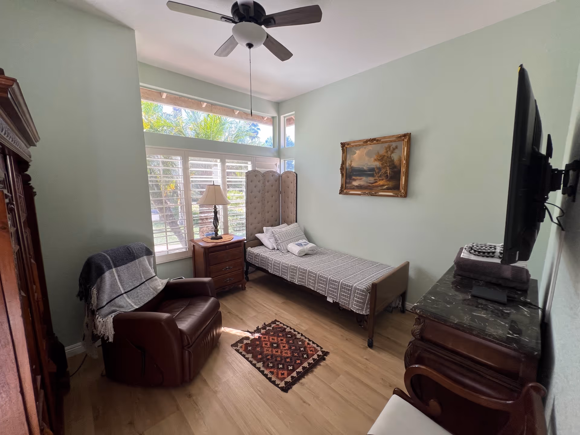 A small, cozy bedroom with a single bed covered in a checkered bedspread, a wooden nightstand with a lamp, a brown leather recliner chair with a blanket draped over it, a wall-mounted flat-screen TV, a decorative folding screen near the window, and a framed painting on the light green wall. The room has large windows with white shutters letting in natural light and a ceiling fan above.