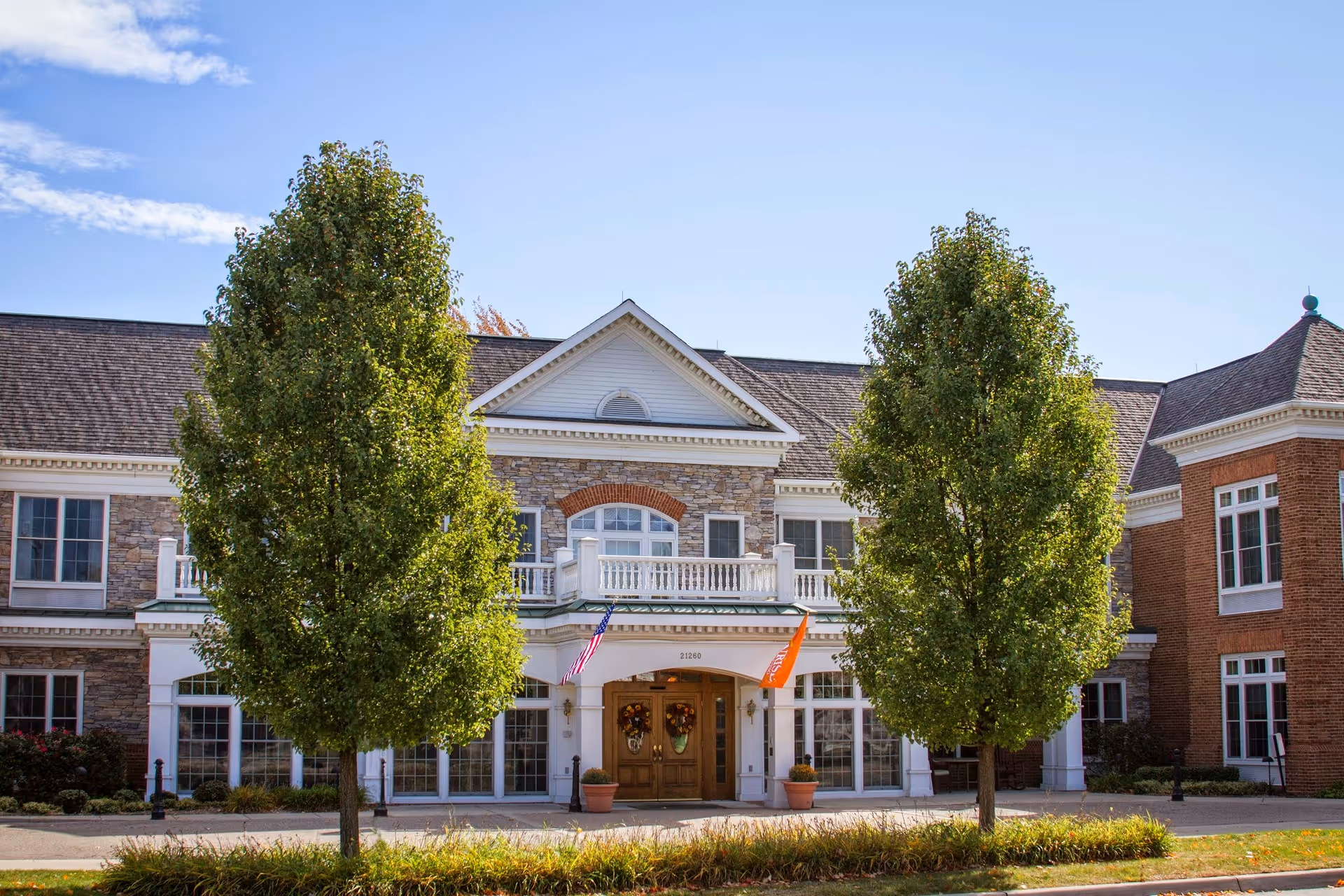Front exterior view of a senior living facility building with a stone and brick facade, two large green trees in front, an American flag and an orange flag near the entrance, and a clear blue sky above.
