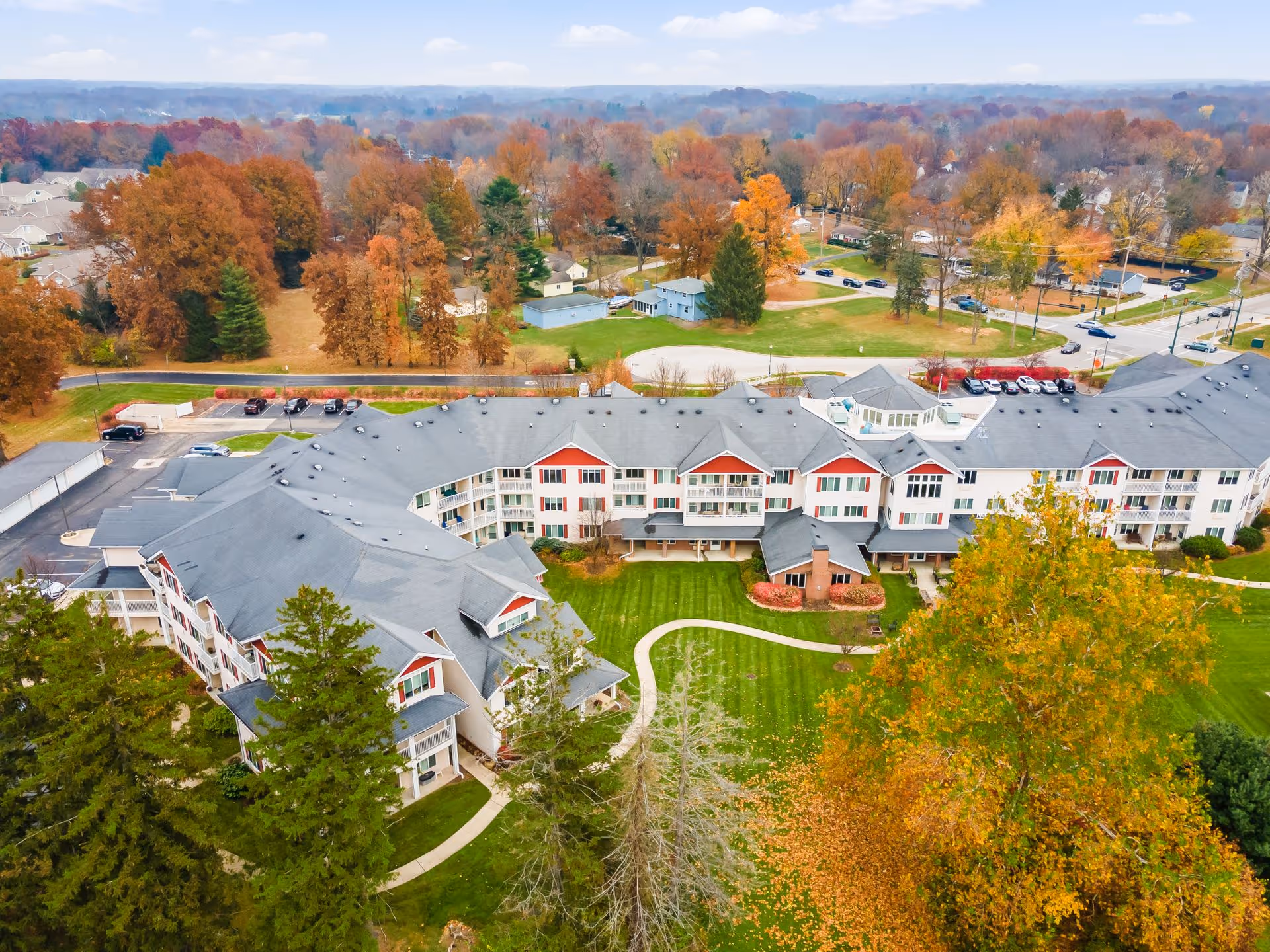 Aerial view of Rittenhouse Village Gahanna senior living facility surrounded by trees with autumn foliage. The building is large with multiple stories, gray roofs, and white walls with red accents. There is a well-maintained green lawn with a winding pathway in front of the building. Cars are parked in the parking lot behind the building, and residential houses and trees are visible in the background.