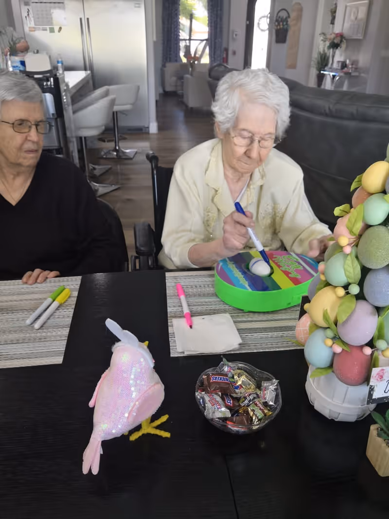 Two elderly women sitting at a table in a living room or common area. One woman is painting an Easter egg with a marker, while the other watches. On the table are colorful markers, a bowl of assorted candy, a decorative Easter egg tree, and a pink plush bird. The background shows a kitchen area and a living room with chairs and a window.