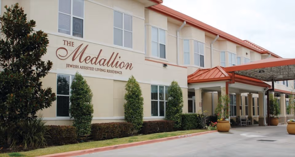 Exterior view of The Medallion Jewish Assisted Living Residence building with beige walls, multiple windows, a red metal roof over the entrance, and landscaping including bushes and small trees along the front.