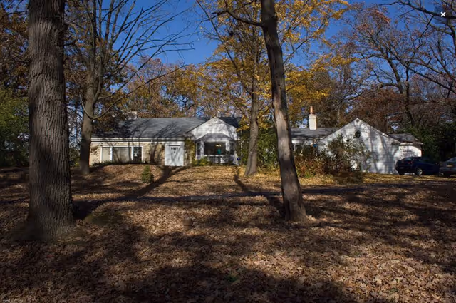 A single-story house surrounded by tall trees with autumn leaves covering the ground. The house has a light-colored exterior with a gray roof and a chimney. There are two cars parked on the right side near the house.