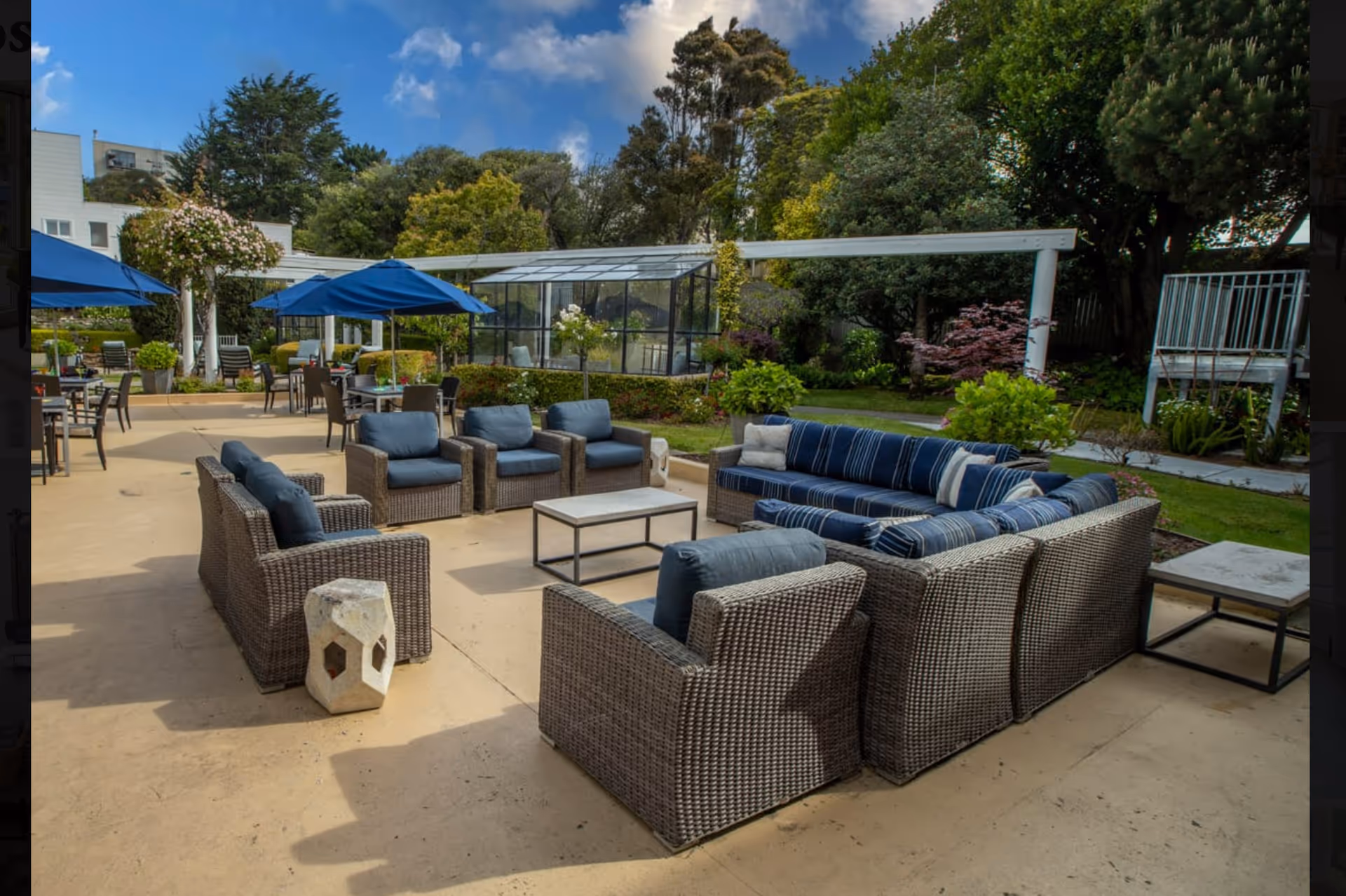 Outdoor patio area with wicker furniture including cushioned chairs and a sectional sofa arranged around a coffee table. Blue umbrellas provide shade over additional tables and chairs. The area is surrounded by greenery, trees, and a glass greenhouse structure under a partly cloudy blue sky.