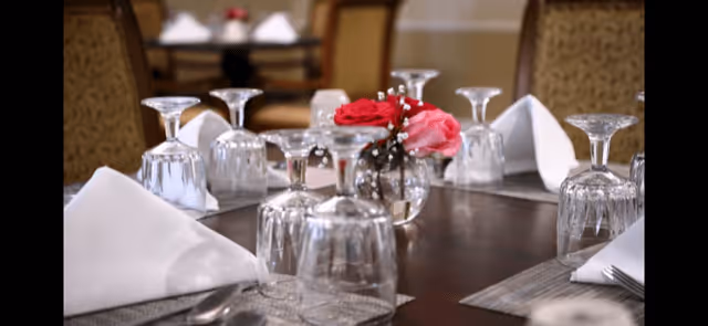 A close-up view of a dining table set with upside-down wine glasses, white folded napkins, and a small vase with red and pink flowers in the center. The background shows more tables and chairs in a softly lit dining area.