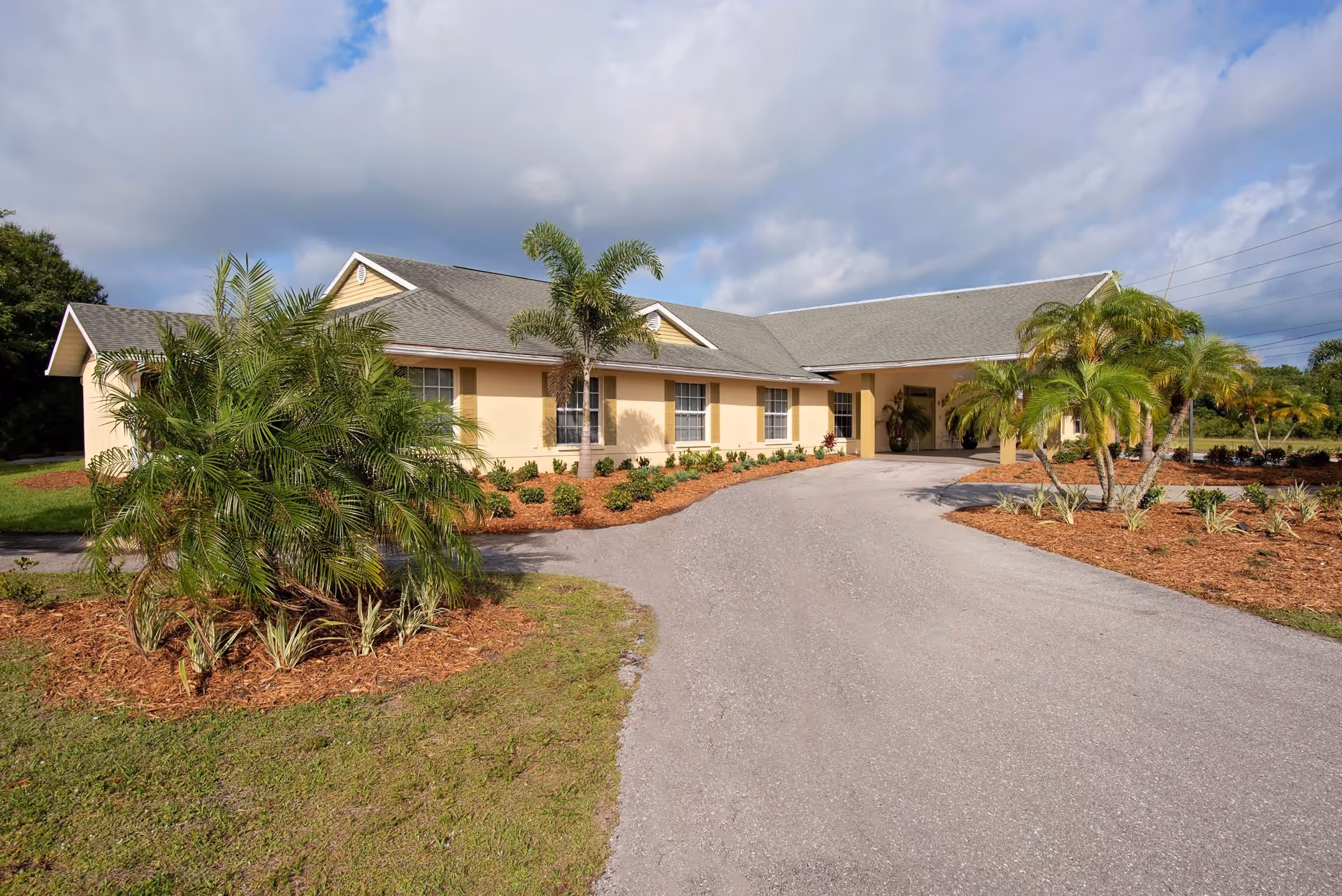 Exterior view of a single-story beige building with a gray roof, surrounded by landscaped greenery including palm trees and shrubs, under a partly cloudy sky.
