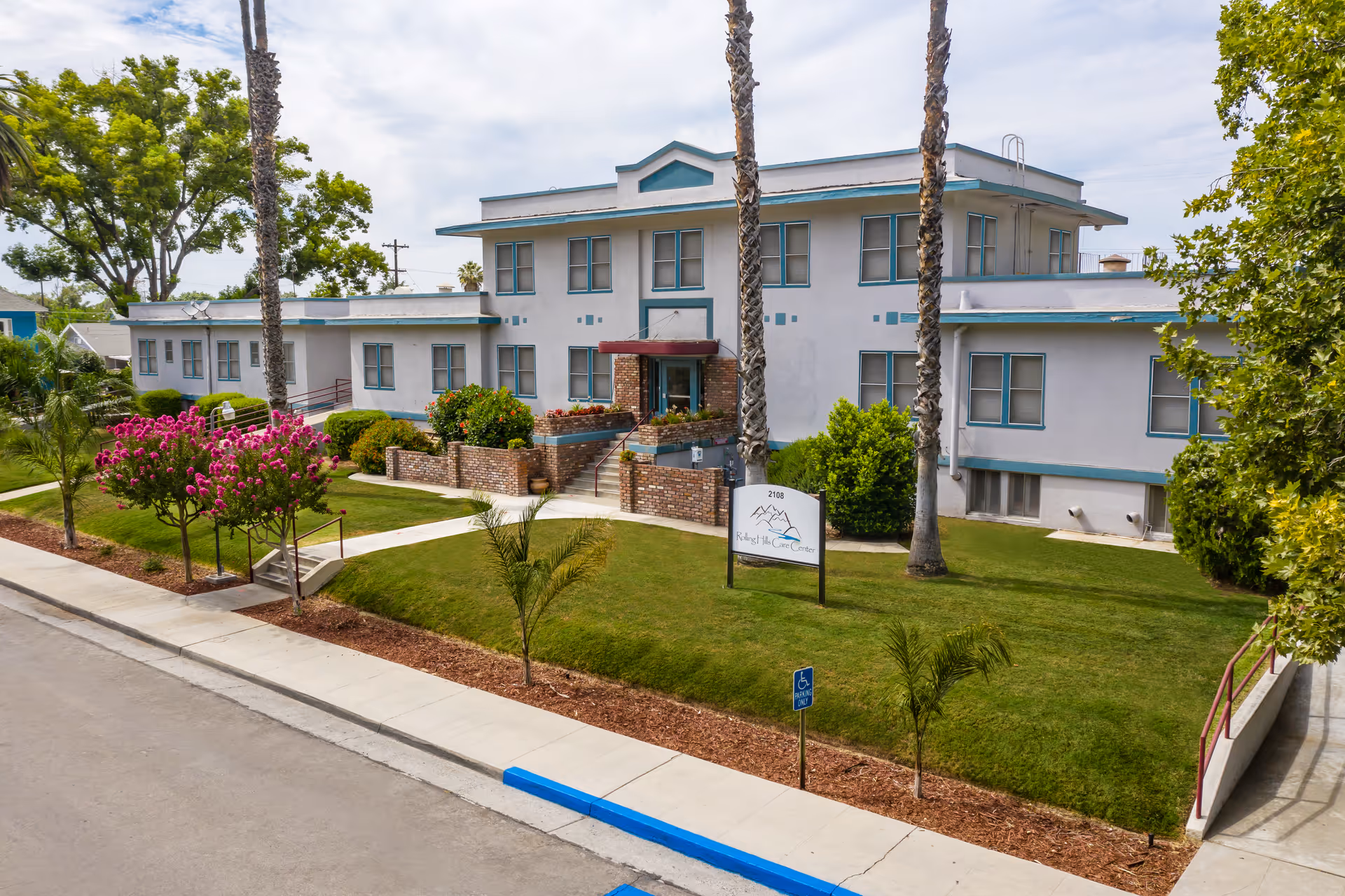 Exterior view of a two-story white building with teal trim, surrounded by a well-maintained lawn, palm trees, and flowering bushes. A sign in front reads 'Rolling Hills Care Center'. The building has multiple windows and a brick entrance with stairs and a ramp.