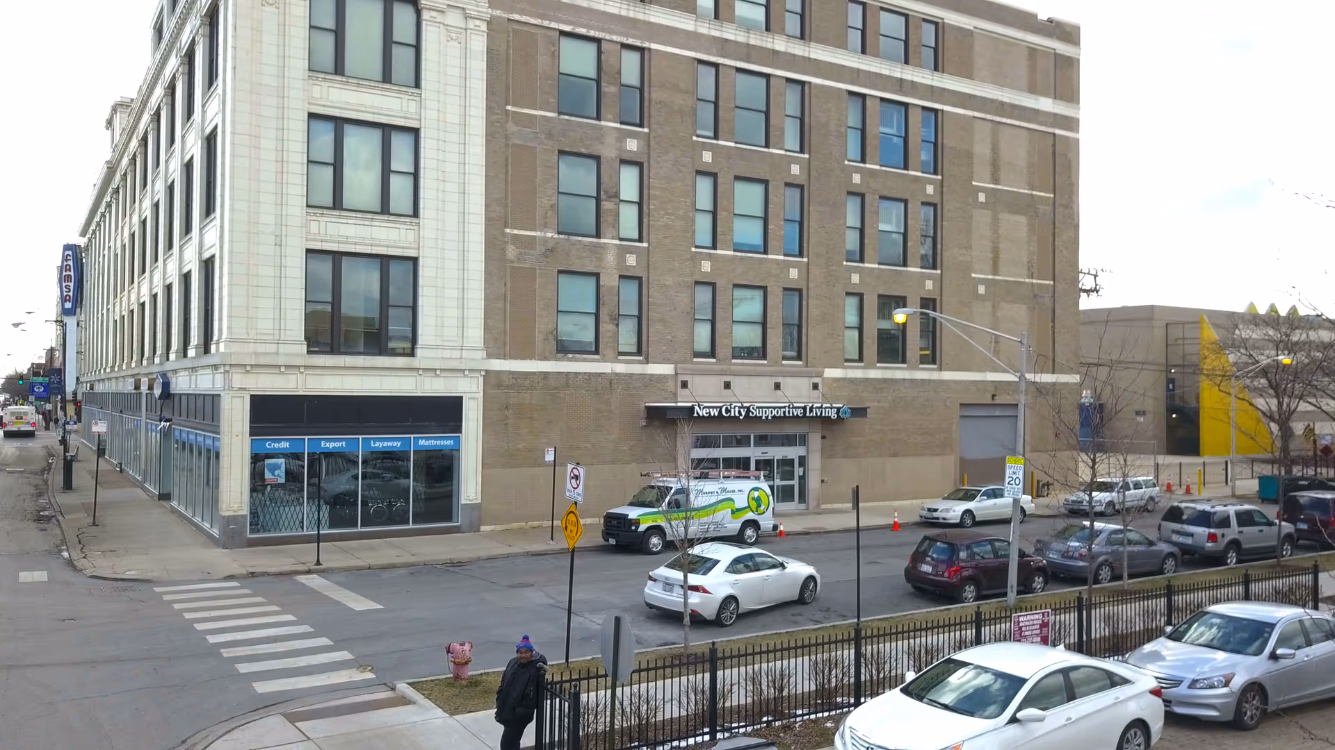 Exterior view of a multi-story brick and stone building on a city street corner with parked cars along the street. The building has a sign above the entrance that reads 'New City Supportive Living'. There is a pedestrian crosswalk and a person walking on the sidewalk near the corner.