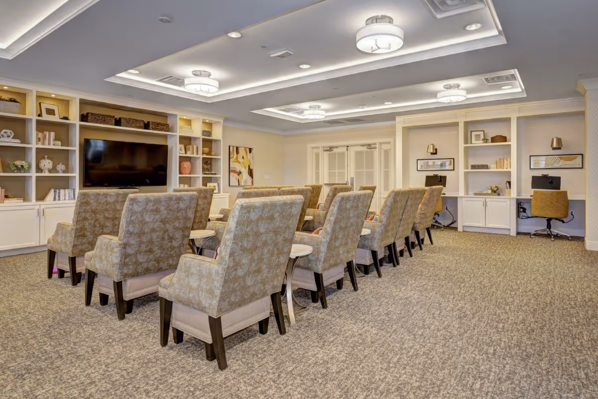 A well-lit common area with rows of upholstered chairs facing a large flat-screen TV mounted on a built-in shelving unit. The room features carpeted flooring, recessed ceiling lights, and built-in desks with chairs along the right wall. Decorative items and framed artwork are displayed on the shelves and walls.