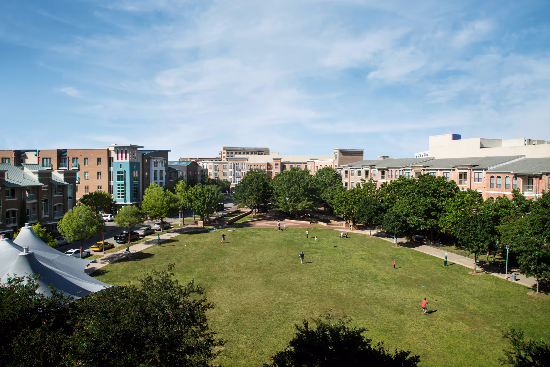 A large green lawn area surrounded by multi-story residential buildings with trees lining the pathways. Several people are scattered across the lawn, enjoying the outdoor space under a partly cloudy blue sky.