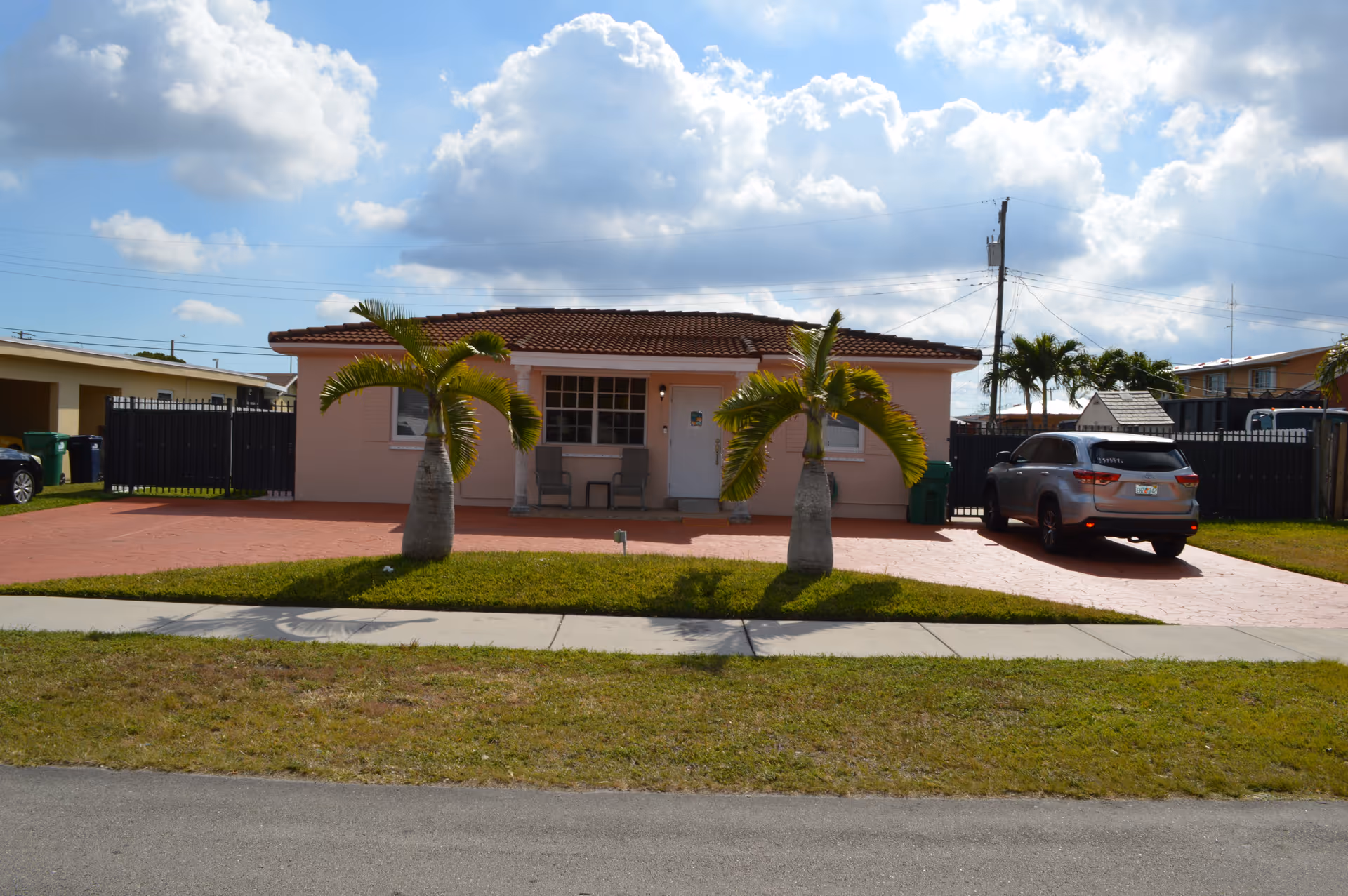 Single-story house with a tiled roof and light pink exterior. Two small palm trees are planted in the front yard with a red paved driveway. There are two chairs on the front porch and a silver SUV parked on the driveway. The sky is partly cloudy.