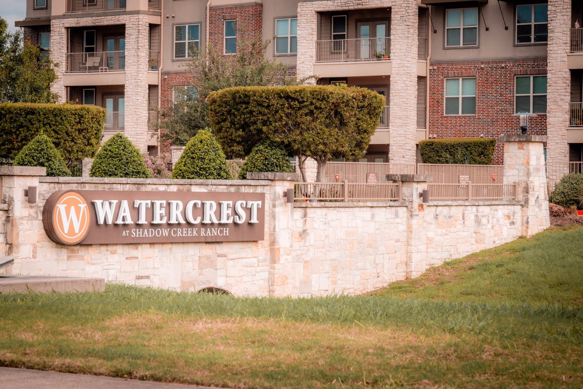 Exterior view of Watercrest at Shadow Creek Ranch senior living facility showing a stone wall with the facility's sign and neatly trimmed bushes in front of a multi-story building with balconies and windows.