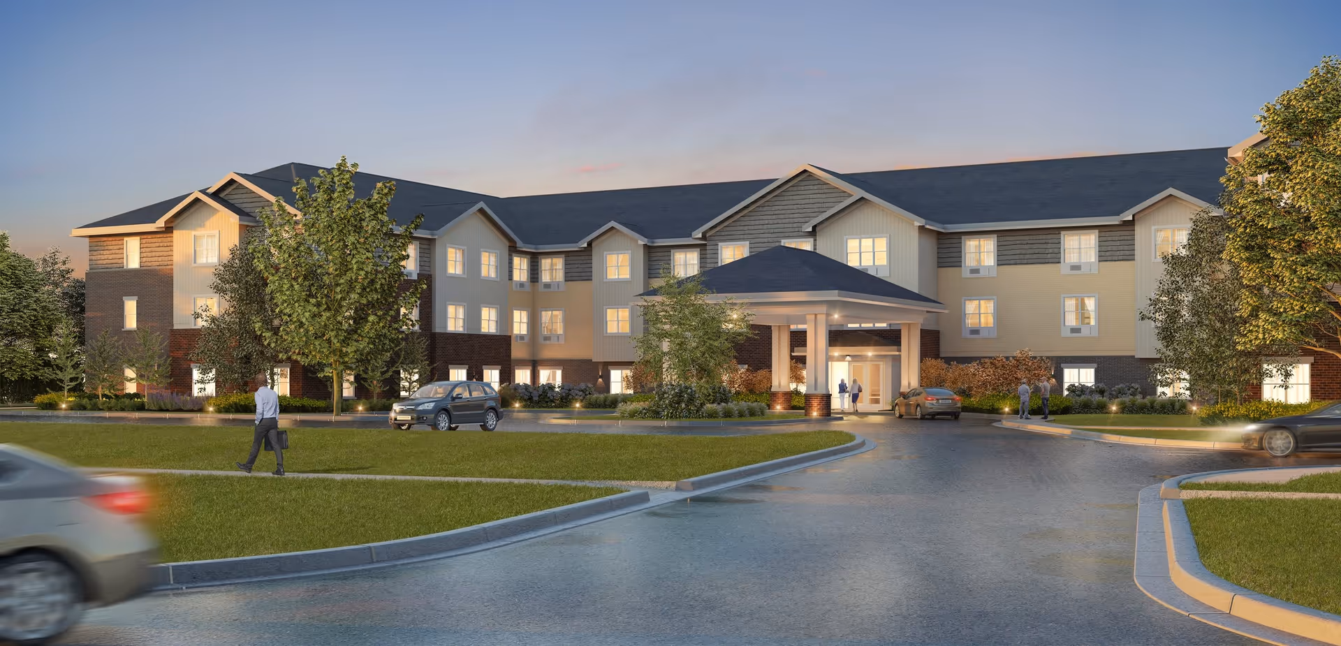 Exterior view of a three-story senior living facility building at dusk with a covered entrance, several cars parked and driving, and a few people walking and standing near the entrance. The building has a combination of brick and siding with multiple windows lit from inside, surrounded by trees and landscaping.