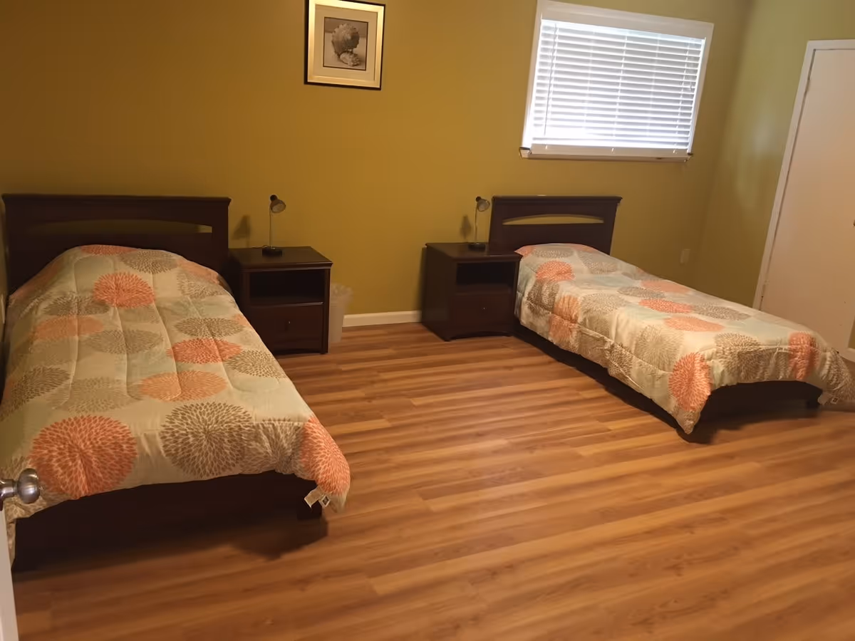 A bedroom with two single beds, each with a floral patterned comforter in shades of orange, beige, and gray. Between the beds are two dark wooden nightstands, each with a small lamp. The room has light wood flooring, a window with white blinds, and a framed picture on the wall above the left bed.