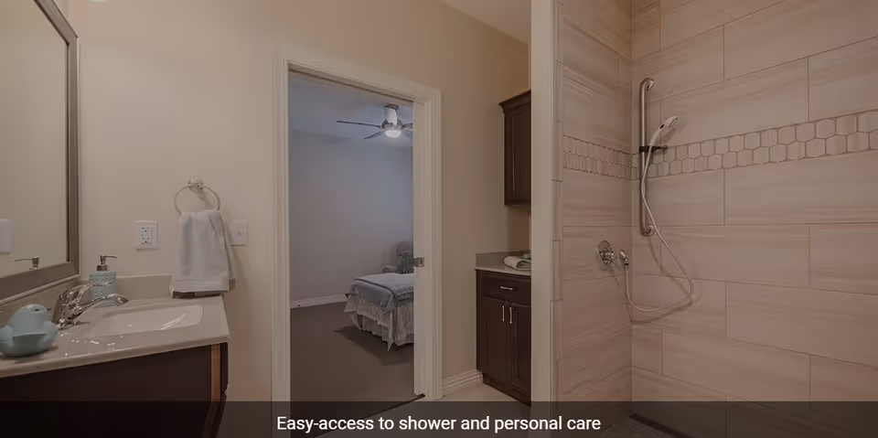 Bathroom with a sink and mirror on the left, an easy-access shower with grab bars on the right, and a doorway leading to a bedroom with a bed and ceiling fan in the background.