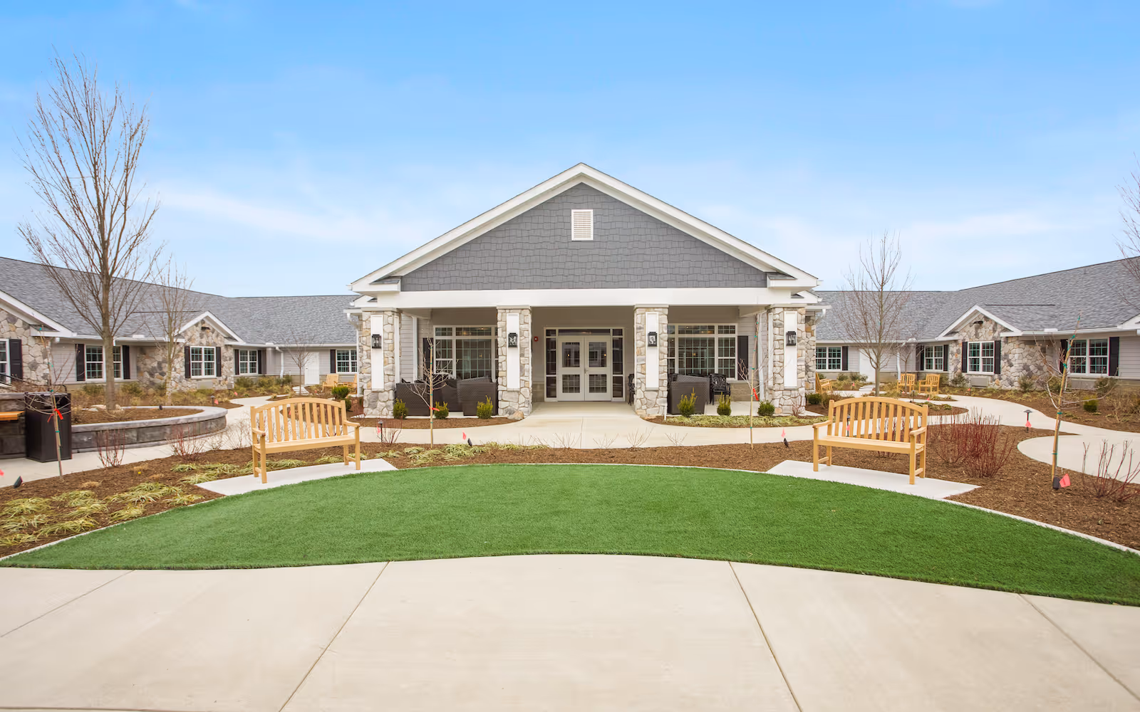 Front exterior view of a single-story senior living facility building with a covered entrance supported by stone pillars. There are wooden benches on either side of a curved walkway, landscaped areas with young trees and shrubs, and a clear blue sky above.