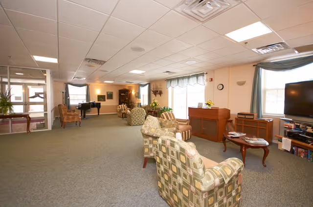 A spacious senior living facility common area with patterned armchairs arranged in a semi-circle, a wooden coffee table, a piano, and large windows with blue curtains allowing natural light to fill the room. The room has a carpeted floor and a ceiling with recessed lighting.