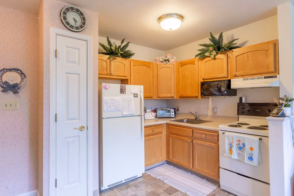 A cozy kitchen with wooden cabinets, a white refrigerator, a white electric stove with a floral towel hanging on the handle, a microwave, a toaster oven, and a sink. There are decorative plants on top of the cabinets and a clock above a white door on the left side. The walls have light wallpaper and the floor has a patterned vinyl covering.