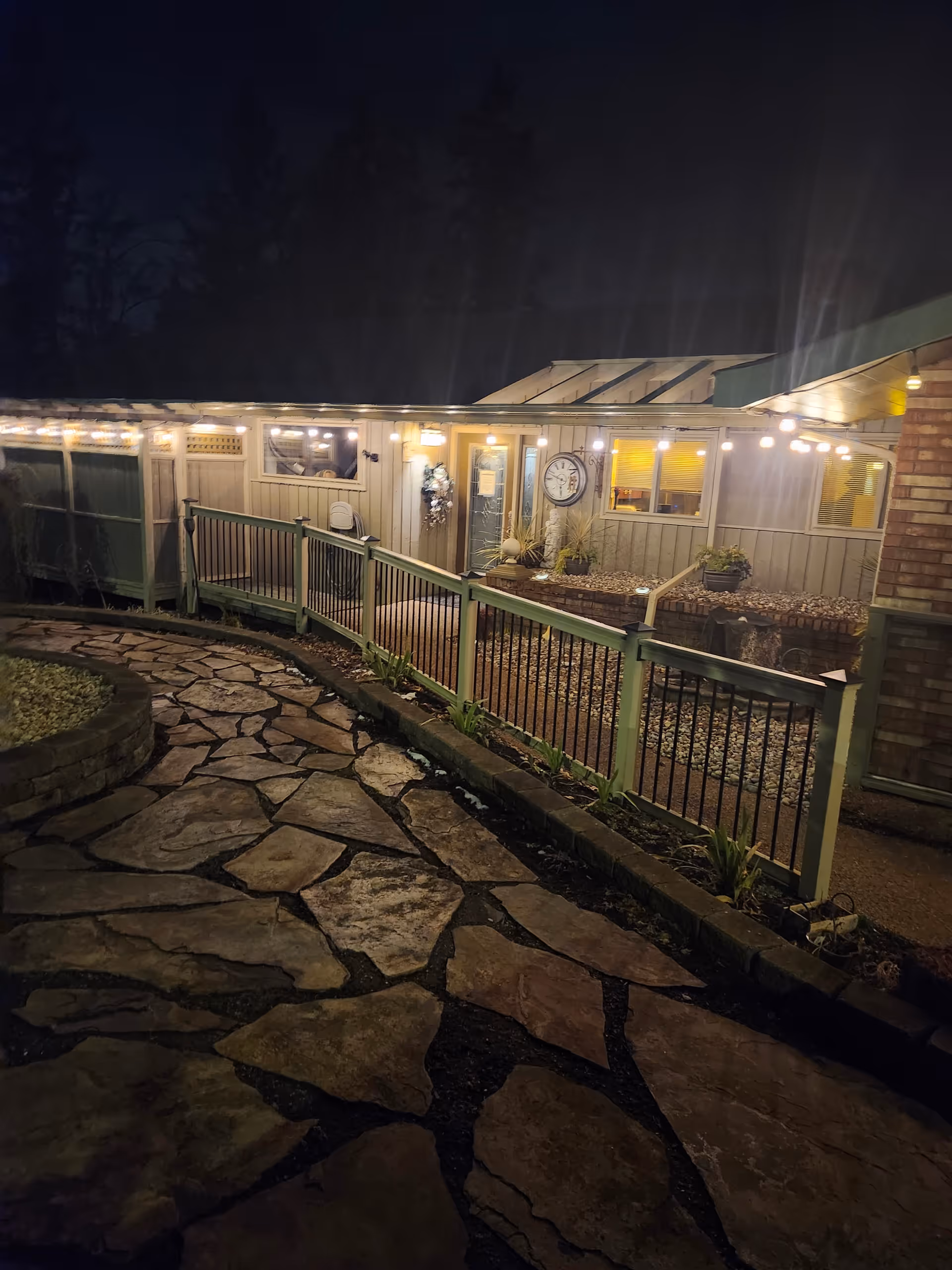Night view of the exterior entrance to Paradise Island Adult Family facility, showing a stone pathway leading to a ramp with railings, string lights hanging above, a clock on the wall, and windows with warm indoor lighting.