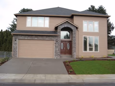 Front exterior view of a two-story house with beige siding and stone accents around the garage and front door. The house has a double garage door, a double front door with glass panels, and multiple windows. There is a concrete driveway and a small lawn with some landscaping.