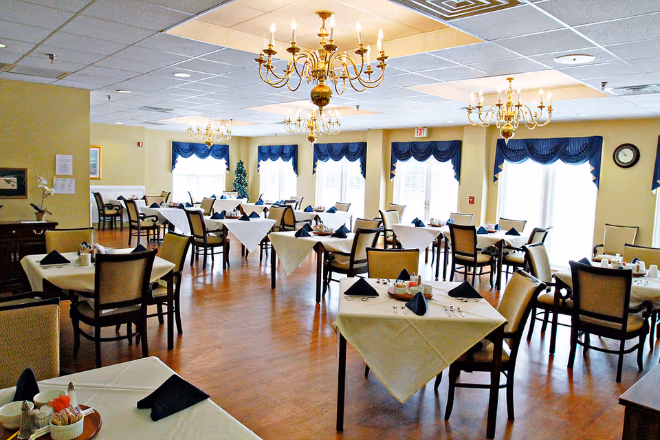 A spacious dining room with multiple tables covered in white tablecloths and set with black napkins, silverware, and condiments. The room features large windows with blue valances, wooden flooring, and several ornate chandeliers hanging from the ceiling. There is a small Christmas tree in the corner and a clock on the wall.