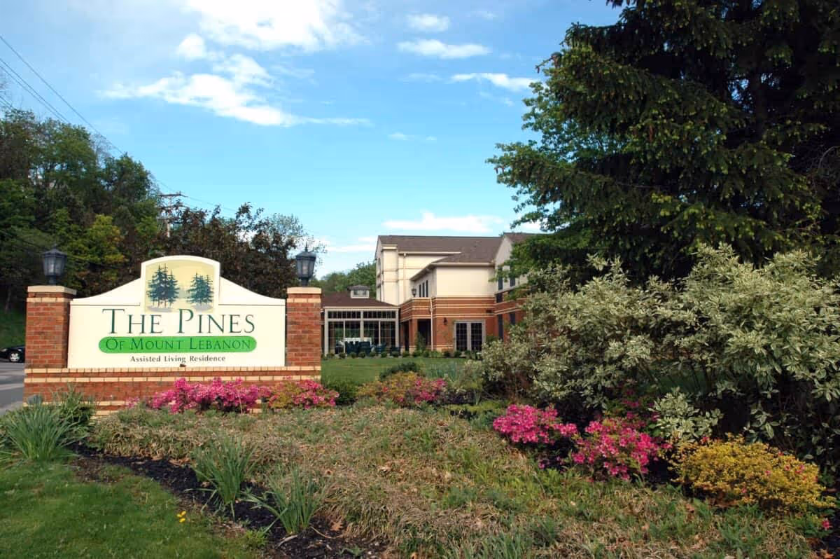 Entrance sign for The Pines of Mount Lebanon assisted living residence surrounded by landscaped greenery and flowers, with the facility building visible in the background under a partly cloudy sky.