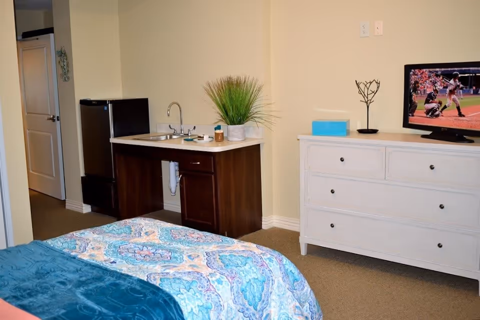 Interior view of a senior living facility room at Danbury Mentor featuring a bed with a colorful patterned bedspread and teal blanket, a small kitchenette with a sink, countertop, and mini refrigerator, a white dresser with a flat-screen TV showing a baseball game, and a decorative plant on the counter.