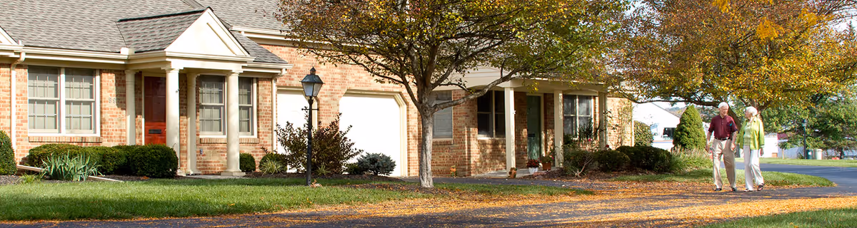 A brick residential building with a front porch, garage, and a tree with autumn leaves in front. Two elderly people are walking on a paved path beside the building under a clear sky.