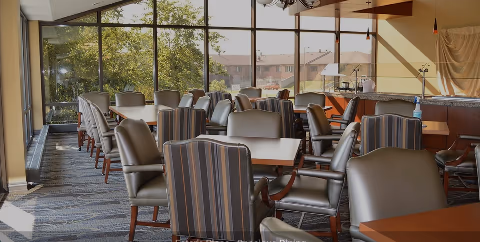 A spacious dining room with multiple tables and cushioned chairs arranged neatly. Large windows on one side allow natural light to fill the room, and outside greenery and buildings are visible through the windows. The room has a carpeted floor and a counter area with a few items on it.