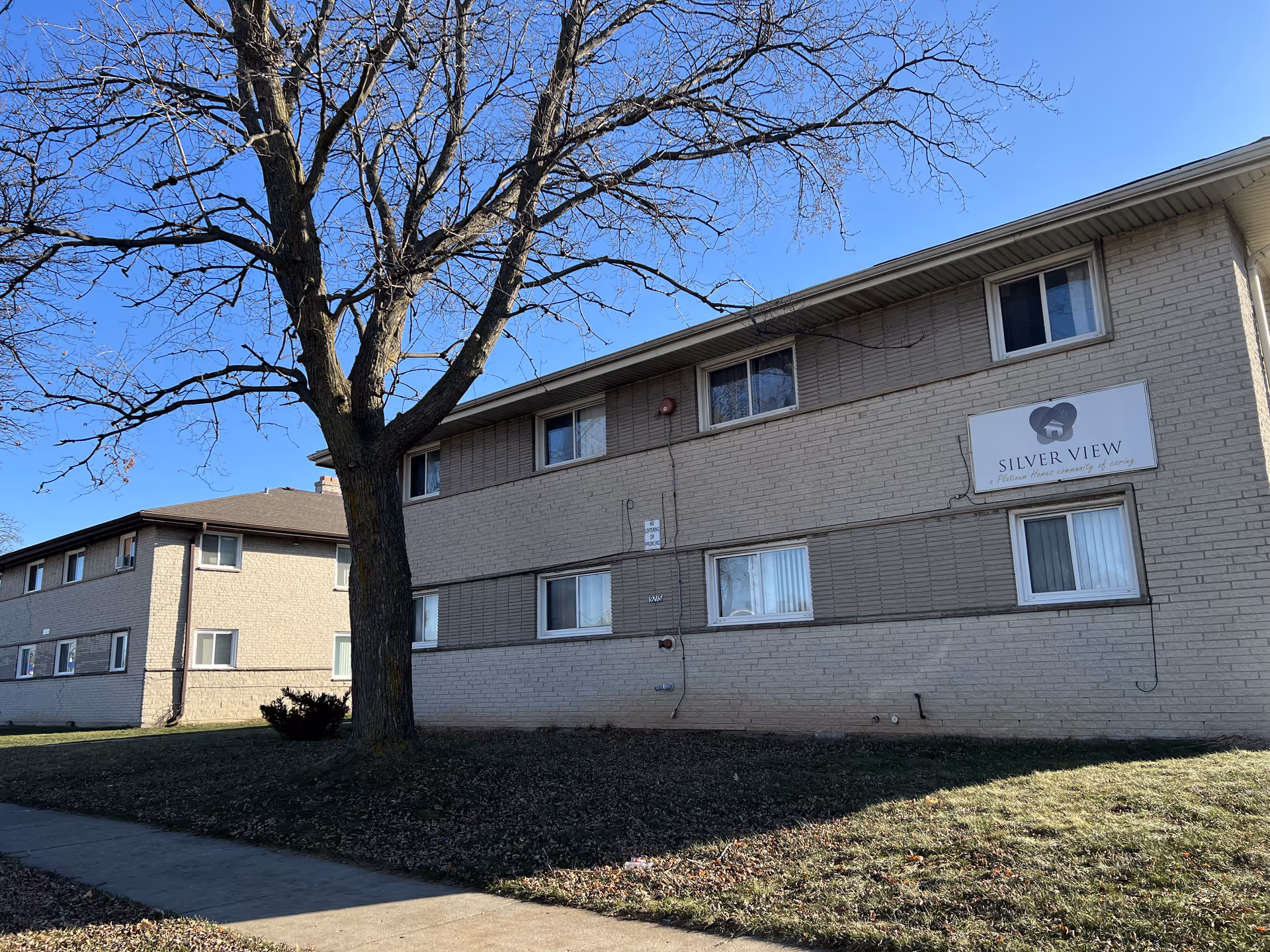 Exterior view of a two-story beige brick building with several windows and a large leafless tree in front. A sign on the building reads 'Silver View' with a heart logo and the tagline 'Precious hours, community of caring.' The sky is clear and blue.