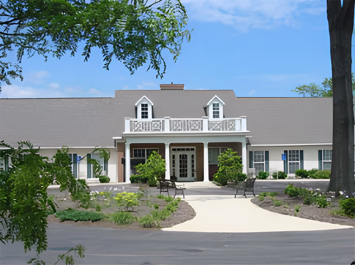 Front exterior view of Abbington of Arlington facility showing a single-story building with a central entrance featuring white columns and a small balcony above. The building is surrounded by landscaped greenery, benches, and a paved walkway leading to the entrance under a clear blue sky.