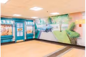 Interior room with a mural painted on the walls depicting a storefront with bread and a street scene including a car and trees. The room has a light wood floor and a drop ceiling with fluorescent lights.