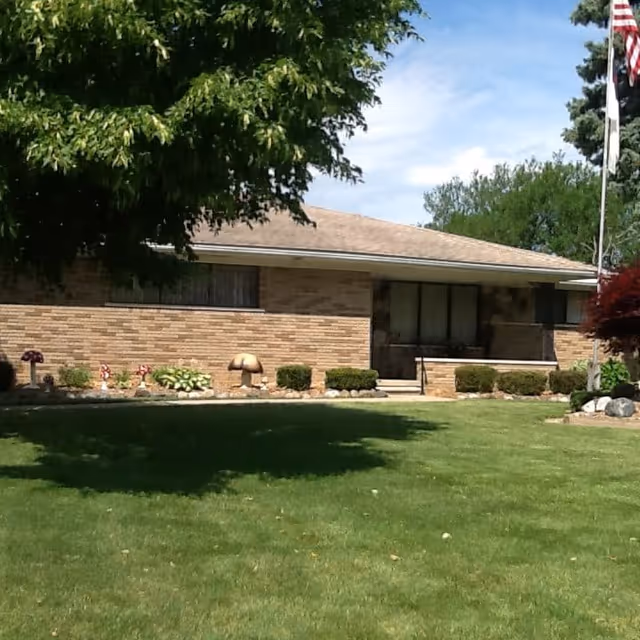 Single-story brick building with a front porch, surrounded by a well-maintained lawn, shrubs, and trees under a partly cloudy sky.