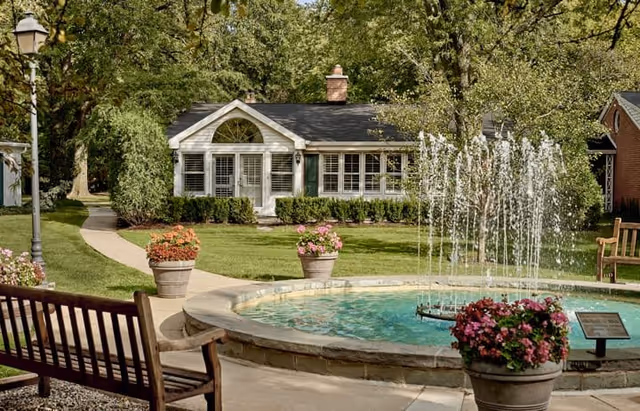 A peaceful outdoor garden area at Westminster Place Independent Living featuring a circular water fountain with multiple streams of water, surrounded by flower pots with colorful blooms. There are wooden benches along a curved pathway, green grass, and trees providing shade. A small white building with large windows and a chimney is visible in the background.