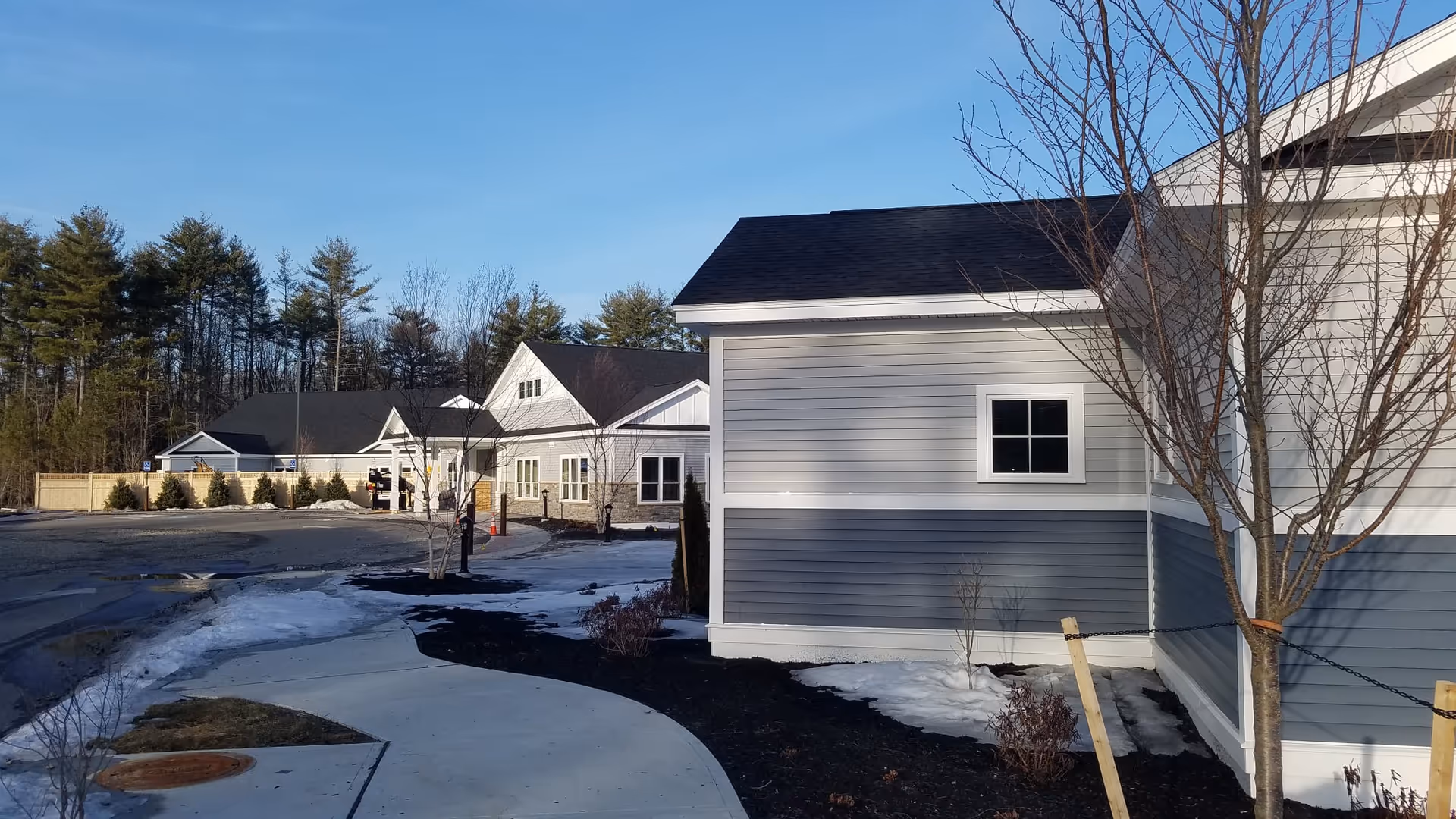 Exterior view of a senior living facility named Summer Commons with multiple connected buildings featuring gray and white siding, black roofs, a curved sidewalk, leafless trees, and patches of snow on the ground under a clear blue sky.
