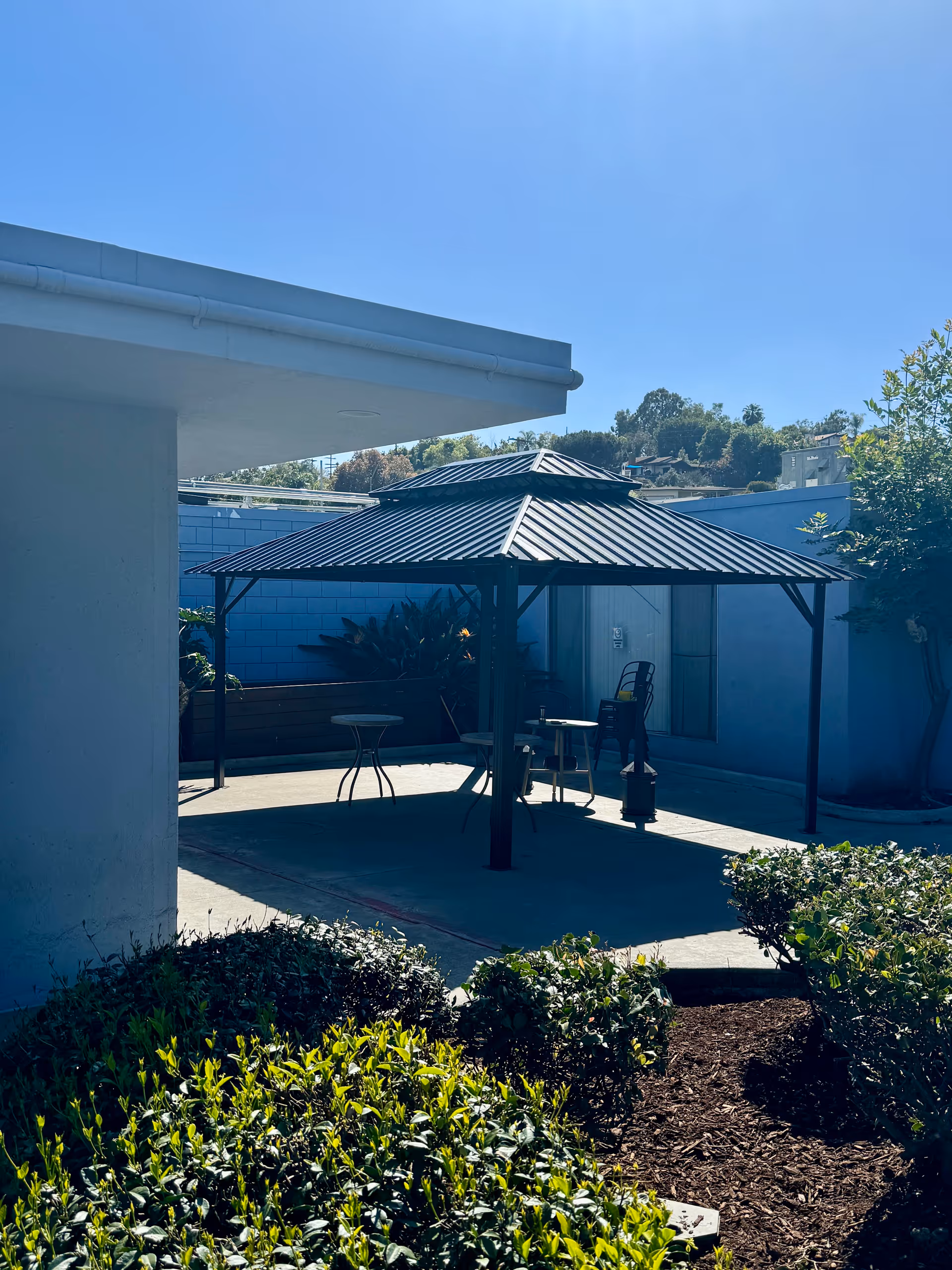 Outdoor patio area with a metal gazebo structure providing shade over several tables and chairs. Surrounding the patio are green bushes and plants, with a clear blue sky overhead.