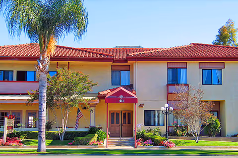 Exterior front view of Fullerton Rosewood Assisted Living facility showing a two-story building with a red tile roof, beige walls, multiple windows, a central entrance with a red awning labeled '411', palm trees, and landscaped greenery with flowers.