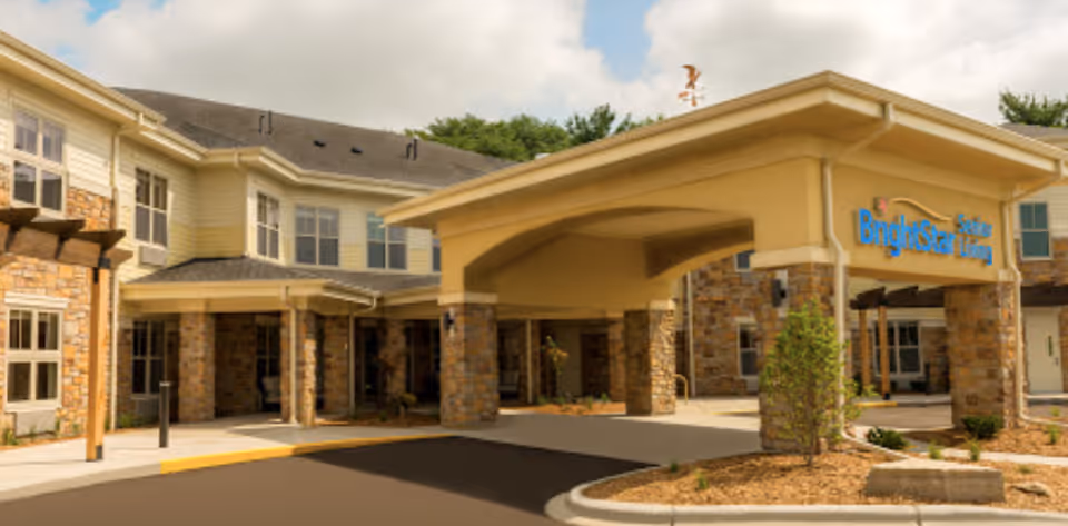Exterior view of Brightstar Senior Living Of Waunakee building entrance with a covered drop-off area supported by stone pillars, surrounded by landscaped plants and trees under a partly cloudy sky.