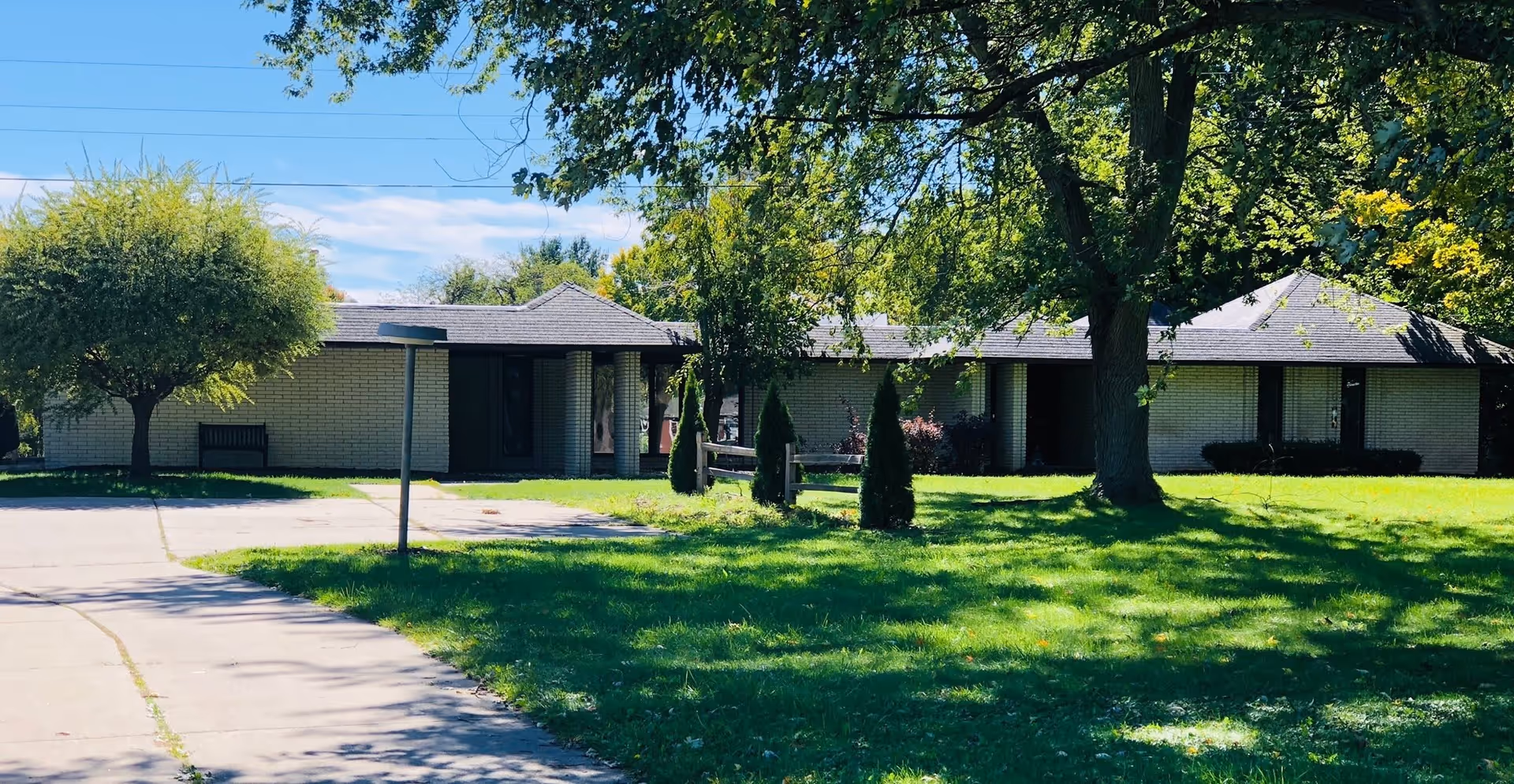 Single-story brick building with a dark roof surrounded by green grass and several trees under a blue sky with some clouds. A paved walkway leads up to the building entrance.